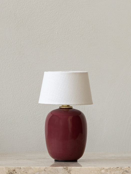 A Torso Portable Table Lamp with a glossy ruby base and a white fabric shade, sitting on marble against a plain wall.