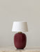 A Torso Portable Table Lamp with a glossy ruby base and a white fabric shade, sitting on marble against a plain wall.