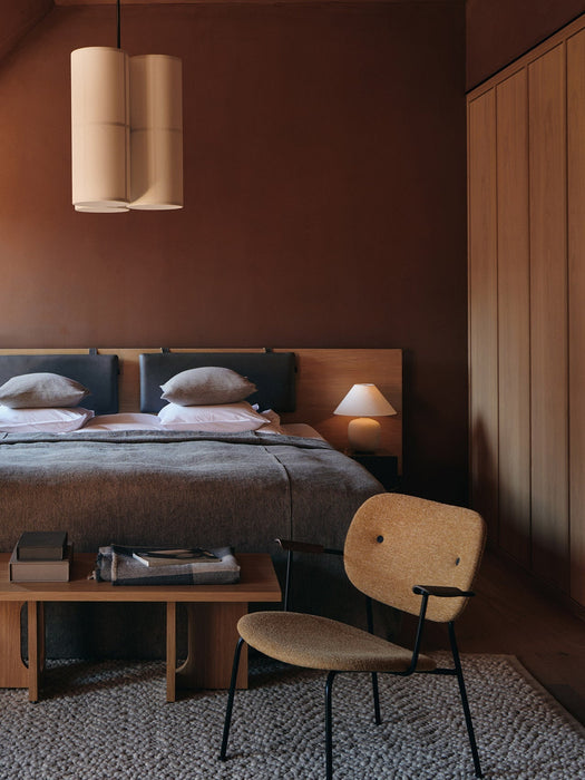 The Torso Table Lamp glowing on a nightstand in a brown-walled bedroom with an Androgyne Lounge Table at the foot of the bed.