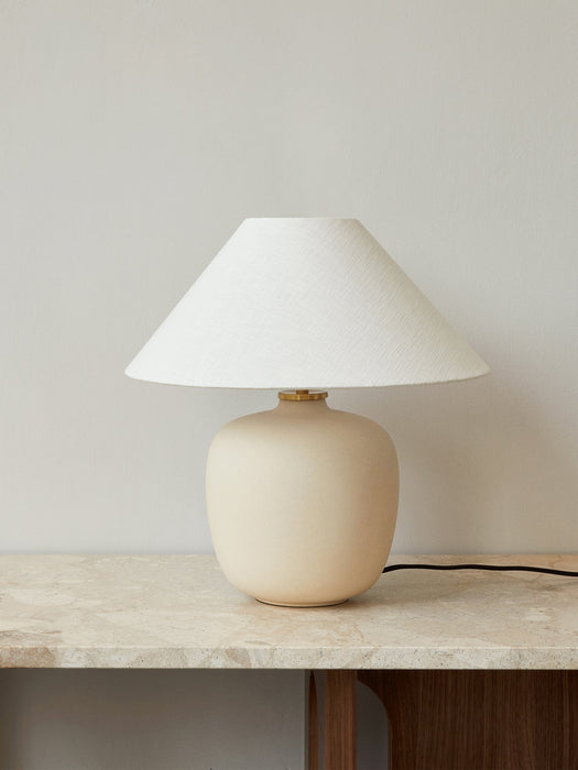 The Torso Table Lamp, 37, with a rounded, sand-coloured base and a conical off-white linen shade, on a marble-topped table.