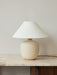 The Torso Table Lamp, 37, with a rounded, sand-coloured base and a conical off-white linen shade, on a marble-topped table.