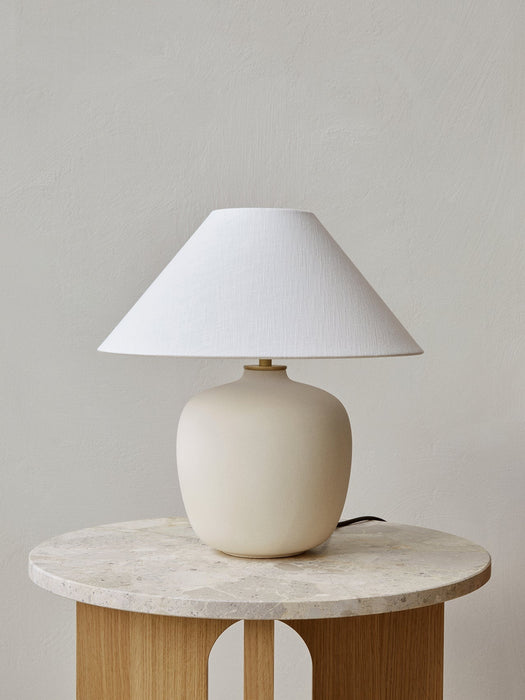 The sand-coloured Torso Table Lamp, 37, with its textured white linen shade, sits on a round marble and wood side table.