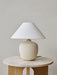 The sand-coloured Torso Table Lamp, 37, with its textured white linen shade, sits on a round marble and wood side table.