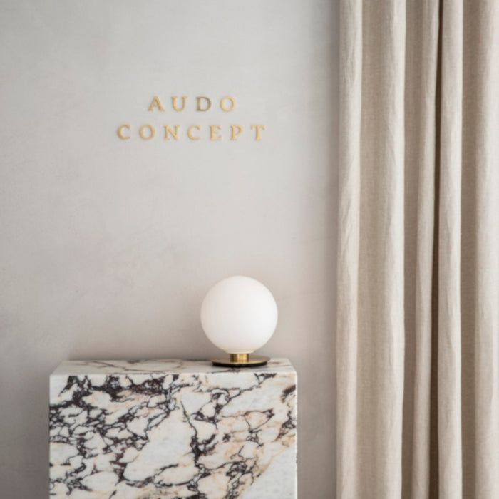 The TR Bulb lamp with its opal glass shade and brass base sits on a veined marble plinth against a light grey wall.