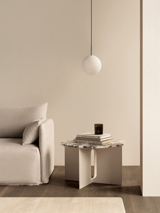 In a minimalist room, a TR Bulb Pendant hangs above an Androgyne Side Table with books, next to a beige Tearoom Sofa.