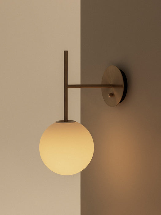 The TR Bulb Suspended Wall Lamp in brushed brass, featuring an opal glass bulb mounted on a corner of a beige and taupe wall.