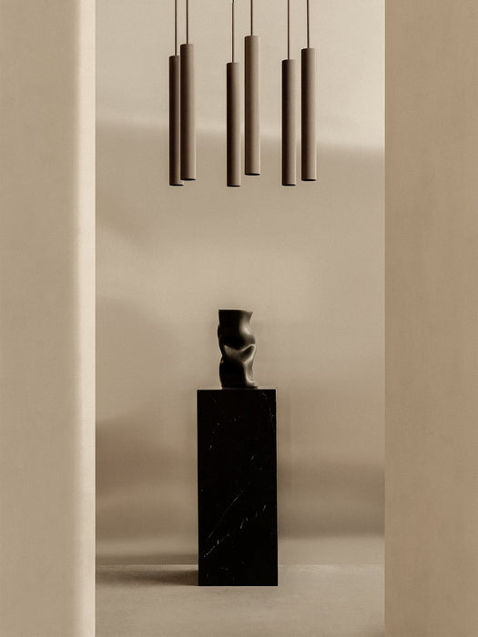 The Tubulaire Chandelier hangs above a black Collapse Vase which sits on a tall black marble Plinth in a minimalist room.