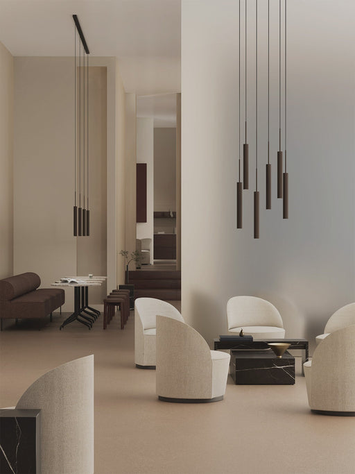 Two Tubulaire Chandeliers hang in a minimalist, modern lounge with beige and brown furniture, armchairs, a sofa, and stools.