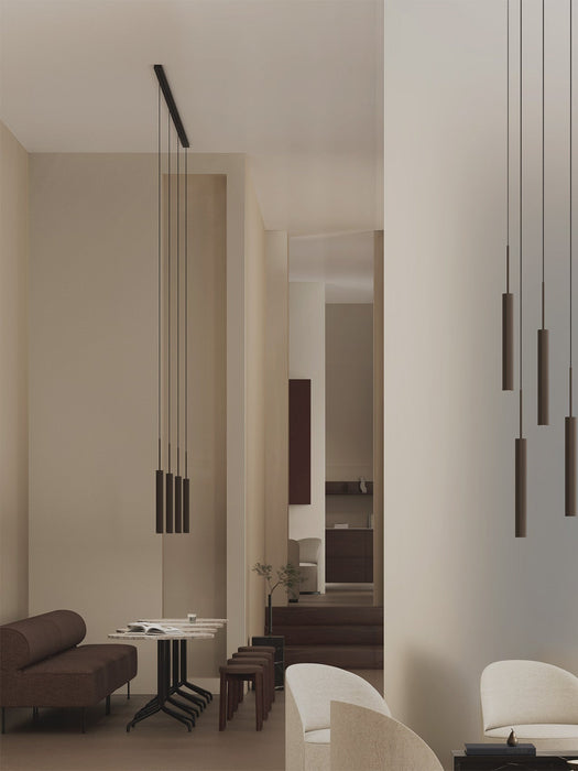 A Tubulaire Rail Pendant illuminates a minimalist dining area with a brown sofa and stools, in a softly lit, neutral room.