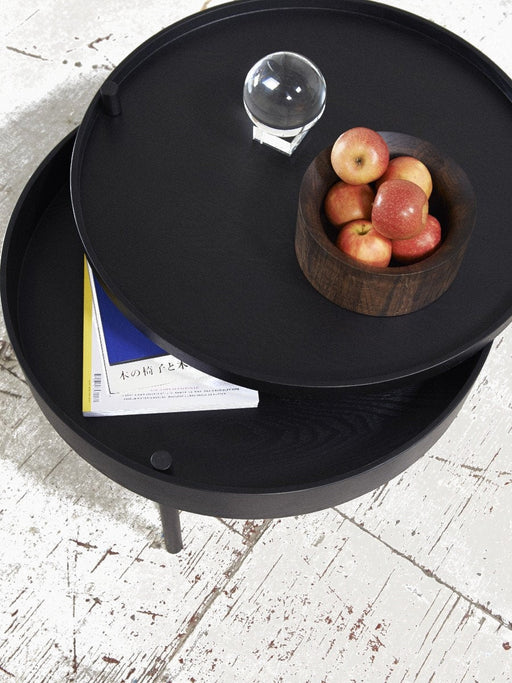 A black Turning Table, its top rotated to show a magazine in storage. On top is a bowl of apples and a glass orb.