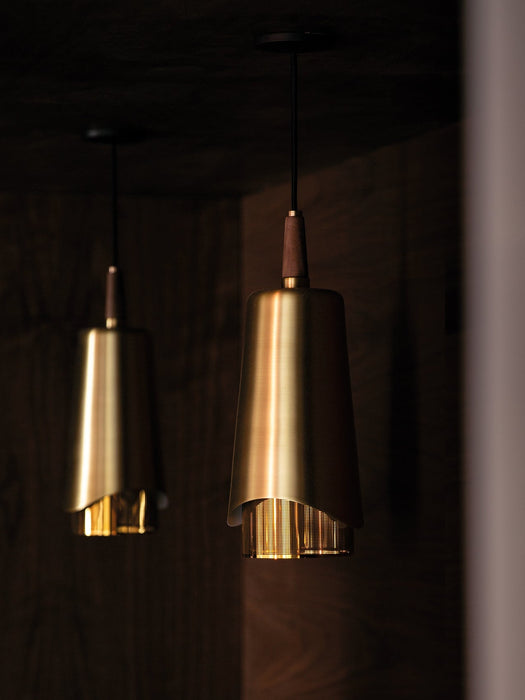 A close-up of two Umanoff Pendants hanging in a dark room, highlighting their warm glowing brass shades and walnut tops.