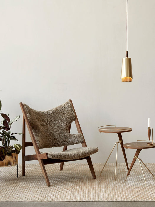 A Umanoff Pendant in brass hangs in a styled room with a wooden lounge chair, two small side tables, and a potted plant.
