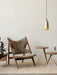 A Umanoff Pendant in brass hangs in a styled room with a wooden lounge chair, two small side tables, and a potted plant.