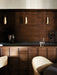 Three brass Umanoff Pendants hang over a dark wood bar counter. On the counter is a wine rack, vase and glassware.