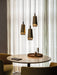 Three lit Umanoff Pendants in brass hang over a round dining table with mustard yellow chairs in front of sheer curtains.