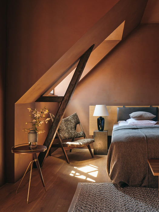 The Umanoff Side Table in a warm, terracotta-toned attic bedroom. The table holds a vase beside a bed and a textured chair.