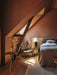 The Umanoff Side Table in a warm, terracotta-toned attic bedroom. The table holds a vase beside a bed and a textured chair.