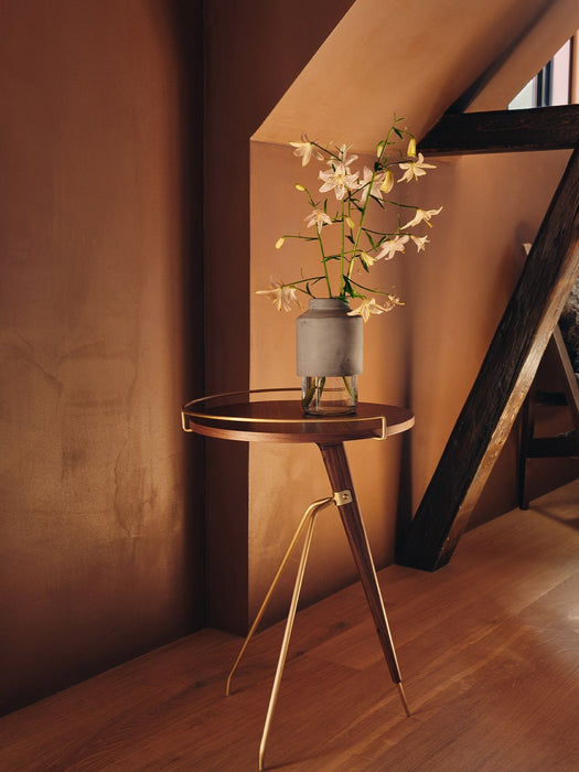 The Umanoff Side Table, with walnut top and brass tripod legs, holds a vase of white lilies in a room with warm, brown walls.