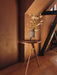 The Umanoff Side Table, with walnut top and brass tripod legs, holds a vase of white lilies in a room with warm, brown walls.