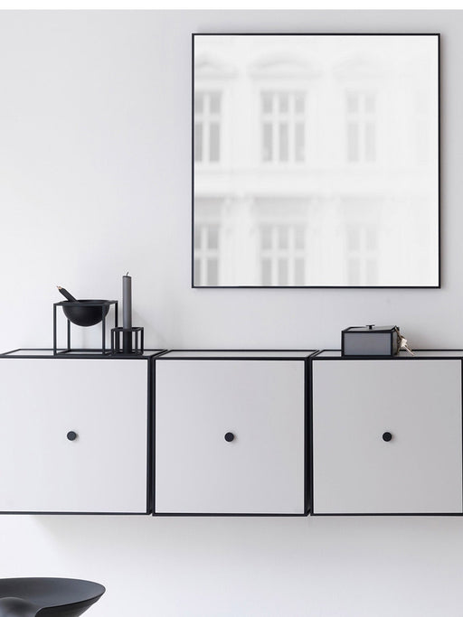 The square, black-framed View Mirror hangs on a white wall above a row of three white cabinets with black frames and knobs.