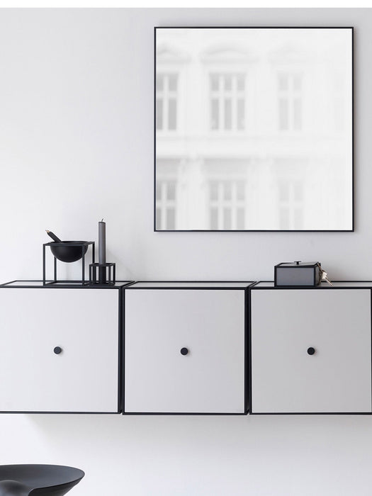 The square, black-framed View Mirror hangs on a white wall above a row of three white cabinets with black frames and knobs.