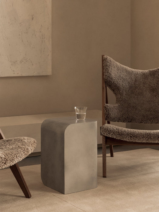 The aluminium Volume Side Table, with a glass of water on top, sits between two sheepskin Knitting Chairs in a beige room.