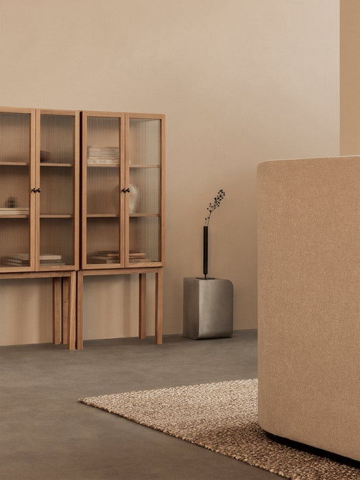 A silver Volume Side Table holding a black vase in a neutral-toned room with a Curiosity Cabinet and beige Tearoom Sofa.