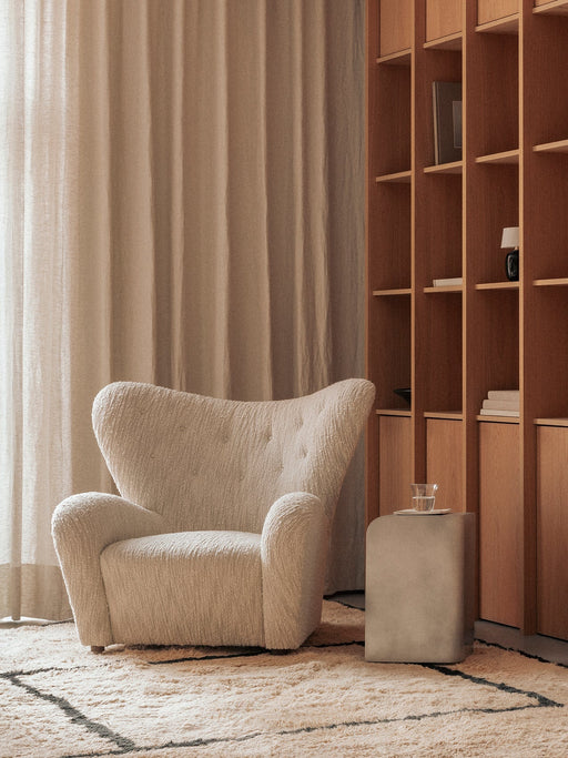 The Volume Side Table in a cozy room, next to a cream boucle Tired Man armchair, with a wooden Curiosity Cabinet in the back.