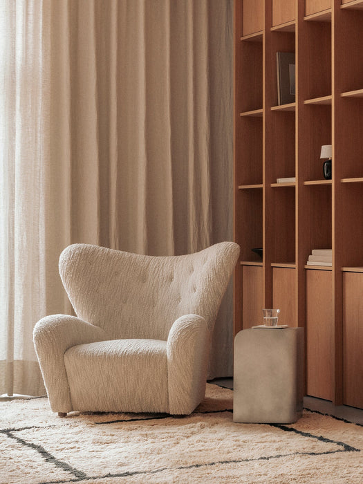 The Volume Side Table in a cozy room, next to a cream boucle Tired Man armchair, with a wooden Curiosity Cabinet in the back.