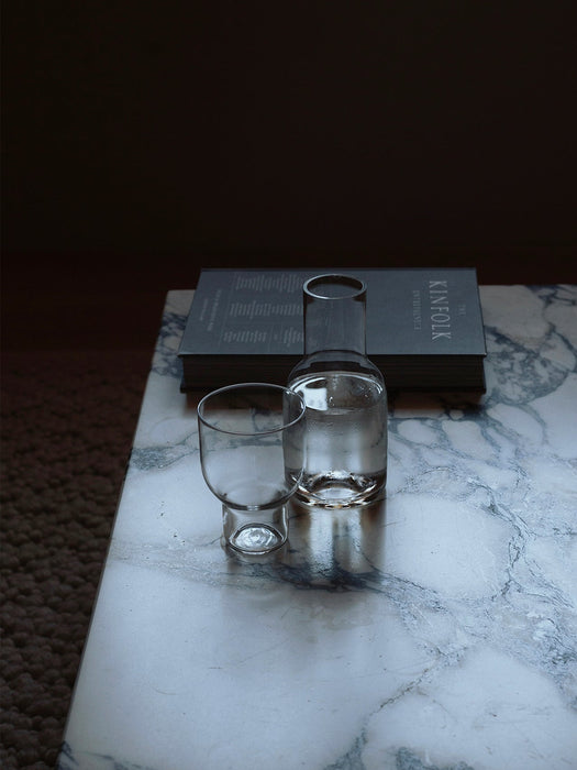 A clear glass carafe with water and matching empty tumbler sitting on a white marble surface, with a book in the background.