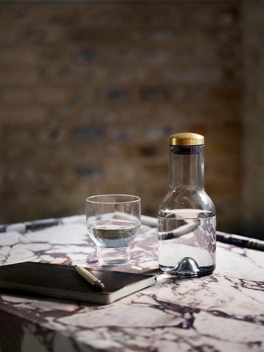 A glass Stackable Carafe with a brass lid and a matching glass, filled with water, sit on a marble table next to a journal.