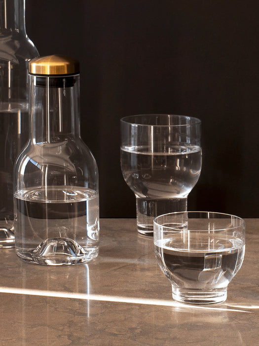 Audo's Bottle Carafe with a brass lid and two glasses by Norm Architects, filled with water on a sunlit stone surface.