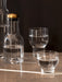 Audo's Bottle Carafe with a brass lid and two glasses by Norm Architects, filled with water on a sunlit stone surface.