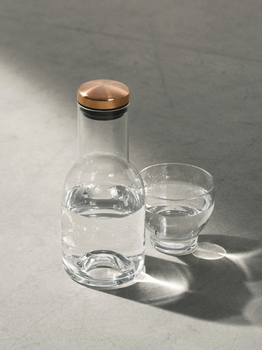 A glass water bottle with a copper lid and an accompanying glass tumbler, both containing water, on a grey concrete surface.