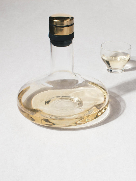 The Audo Wine Breather Deluxe, a wide glass carafe with gold stopper, filled with white wine next to a small drinking glass.
