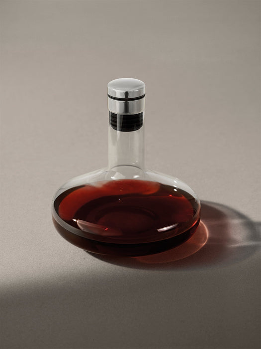 A contemporary glass wine decanter with a wide base, partially filled with red wine, topped with a silver and black stopper.