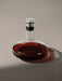 A contemporary glass wine decanter with a wide base, partially filled with red wine, topped with a silver and black stopper.