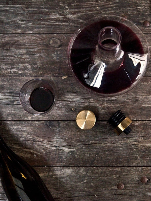 A flat lay of the Wine Breather Carafe with red wine, a glass, its brass stopper, and a bottle on a dark wood table.