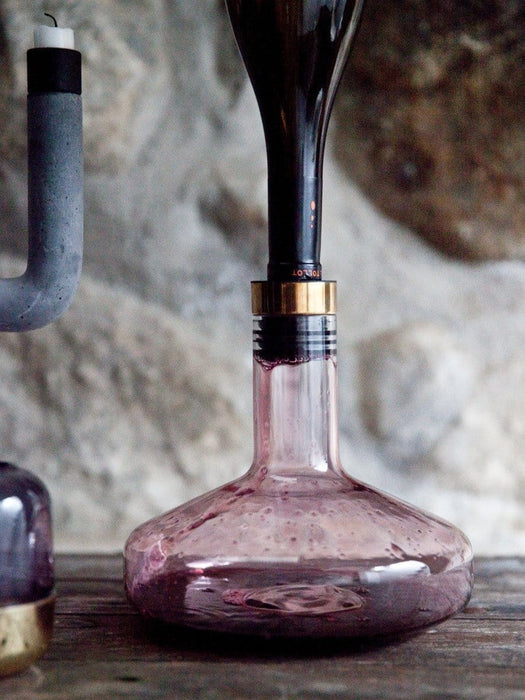 A close-up of the Wine Breather Carafe decanting red wine from an inverted bottle on a wooden table against a stone wall.