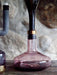 A close-up of the Wine Breather Carafe decanting red wine from an inverted bottle on a wooden table against a stone wall.