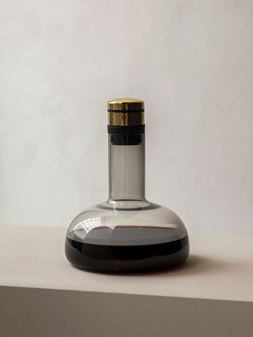 The Audo Wine Breather Carafe in smoked glass with a brass lid, sits on a grey surface, partially filled with red wine.