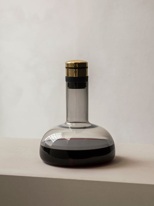 The Audo Wine Breather Carafe in smoked glass with a brass lid, sits on a grey surface, partially filled with red wine.