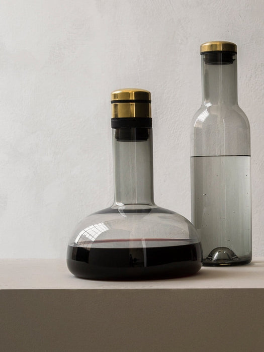 A Wine Breather Carafe of red wine and a Bottle Carafe of water, both in smoked glass with brass lids, on a light surface.