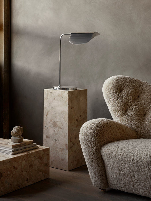 A polished aluminium Wing Table Lamp sits atop a marble Plinth, next to a cream-coloured, textured The Tired Man armchair.