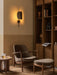 A living room scene with an Elizabeth Lounge Chair, Ottoman, Androgyne Side Table, Tile Rug, and glowing Wing Wall Lamp.