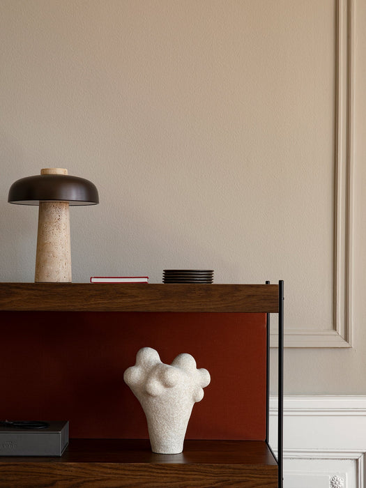 Close-up of a dark wood shelving unit styled with a travertine lamp, coasters, a white abstract vase, and an Audo box.