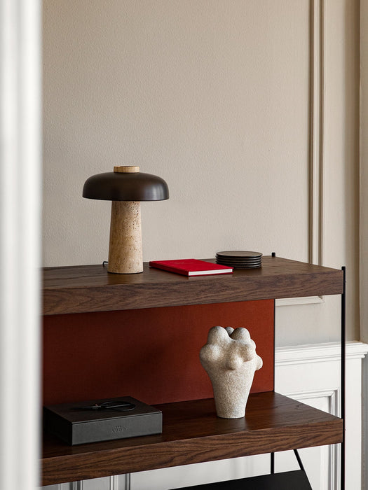 A styled dark wood shelf with a travertine table lamp, red notebook, black leather box, and a textured abstract sculpture.