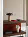 A styled dark wood shelf with a travertine table lamp, red notebook, black leather box, and a textured abstract sculpture.