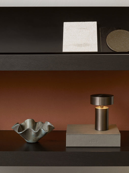 A lit bronze Column Table Lamp and a wavy ceramic bowl sit on a dark wood shelf. Above, another shelf displays samples.
