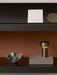 A lit bronze Column Table Lamp and a wavy ceramic bowl sit on a dark wood shelf. Above, another shelf displays samples.